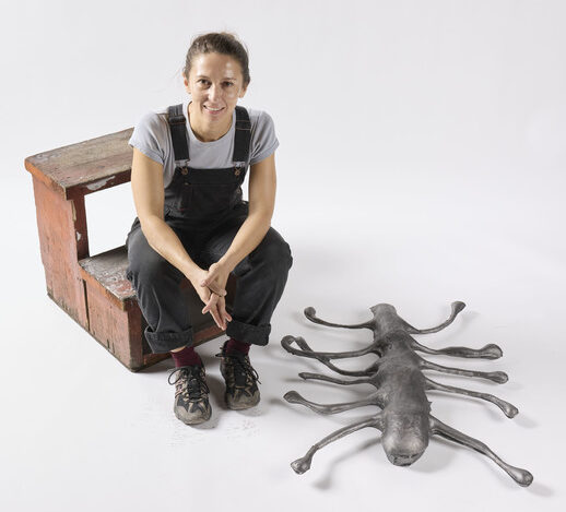 Description: A woman wearing overalls sits on a wooden step stool in a grey space. Next to her is a cast-metal sculpture that looks like an insect with many long arms.